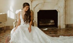 A bride sitting in a vintage chair, wearing a detailed white wedding dress with a long train in a room with a rustic fireplace