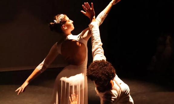 Two dancers, a man and a woman, wearing white, strike a post on a dark stage