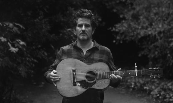 Matt Nathanson poses with his guitar in front of trees and bushes. 