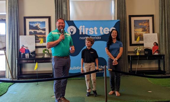 Three people stand in front of a display about First Tee, a man in a green shirt and two young people in blue shirts