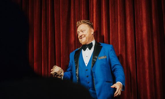 A magician, Bill Abbott, interacts with his audience while standing in front of a red velvet curtain