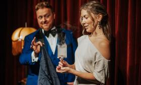 A magician in a blue tux, conjures magic with an audience member in off white holding a smoking glass