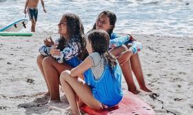 Girls at Surf Camp, sitting on a board in the sand as they listen to instruction