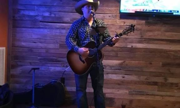 Lane Maury clutches his guitar and performs in front of a wall with wooden panels. 