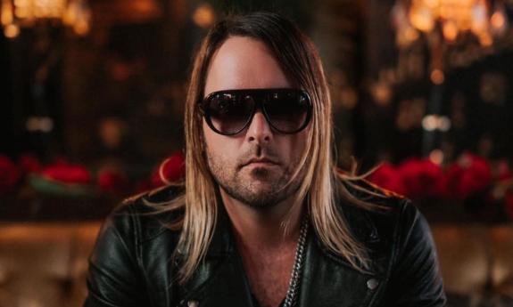 Clayton Sturgeon wears black glasses while posing in front of a blurred backdrop. 