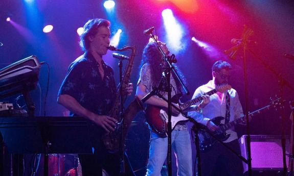 Three musicians from the band Star Collar (saxophone, bass, guitar) on a stage beneath multi color lights.