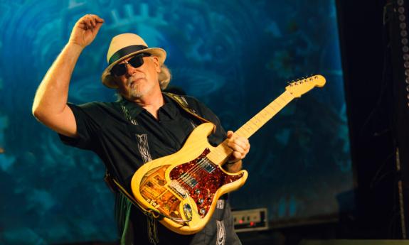 Tas Cru strums his guitar while wearing a hat and glasses. 