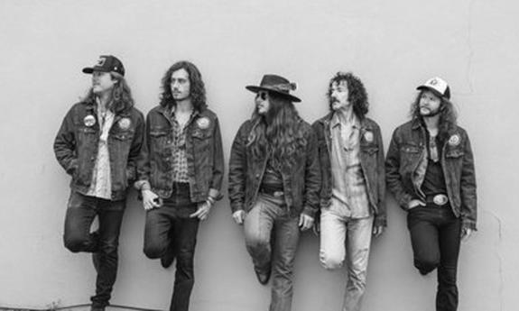 The band Hippies & Cowboys standing against a white wall, each with a foot on wall