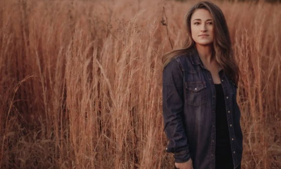 Jennifer Knapp stands against a backdrop of wheat, wearing a black jacket. 