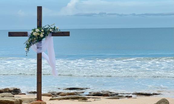A cross on the beach, decorated for a beach wedding with roses and greenery and a white veil