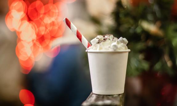A cup of hot cocoa mounded with marshmellows, sitting in front of red holiday lights