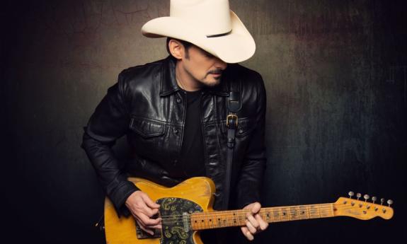 Brad Paisley wears a white cowboy hat and leather jacket while clutching his guitar in front of a black backdrop. 