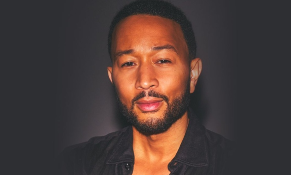 John Legend wears a black shirt and poses in front of a black backdrop.