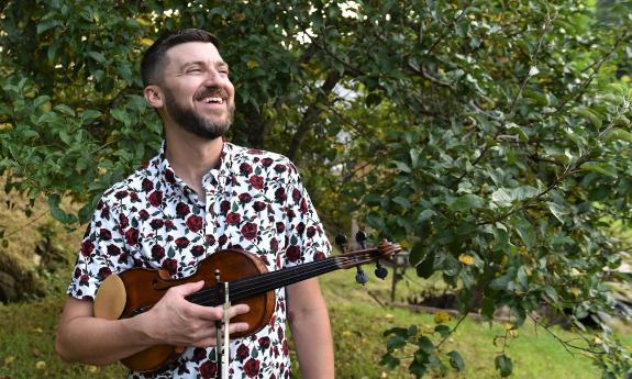 Musician Josh Goforth, outside near a tree, holding his fiddle