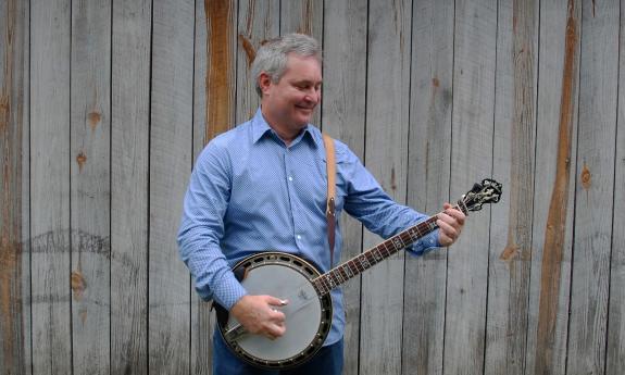 Scott Anderson, wearing a blue shirt and playing banjo in front of a grey board fence