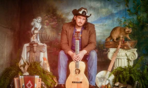 Musician and songwriter Willi Carlisle, seated with props and a colorful backdrop