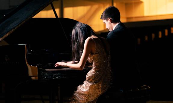 Two piaianists playing the same grand piano, facing away from us