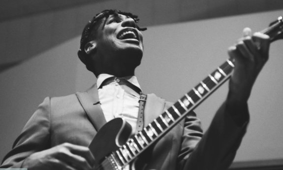 Jon Batiste smiles and stums his guitar.