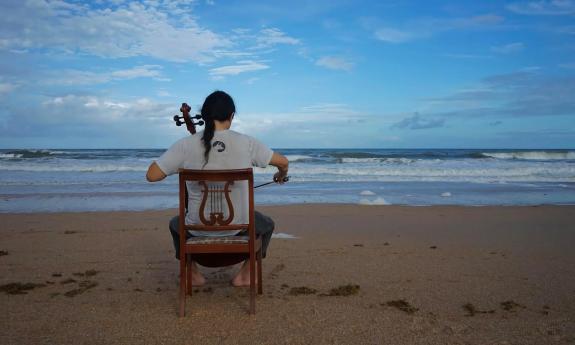 A cellist on a wooden chair, facing the Atlantic Ocean while he plays the cello