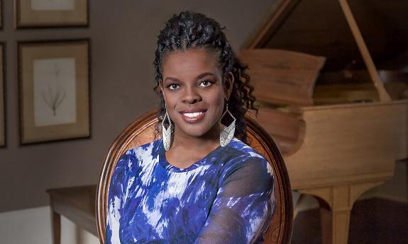 Soprano Ann Marie McPhail, wearing a white and blue dress and sitting with a light wood-toned piano behind her