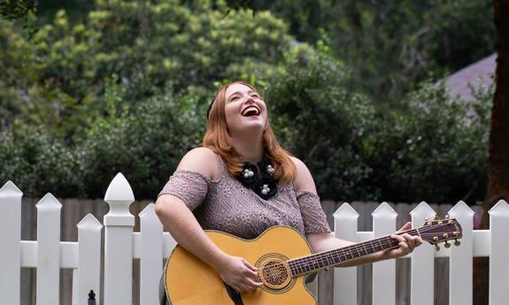 Singer-songwriter Lauren Mullinax with her guitar, laughing in front of a picket fence