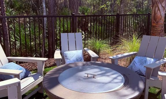 A firepit/table surrounded by comfortable chairs, near a fence with a view of the woods