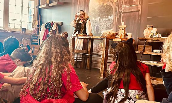 Children sitting in a local museum listening to a woman wearing colonial clothing