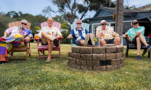 Five colorfully-dressed band members, sitting in Adirondack chairs by a fire pit