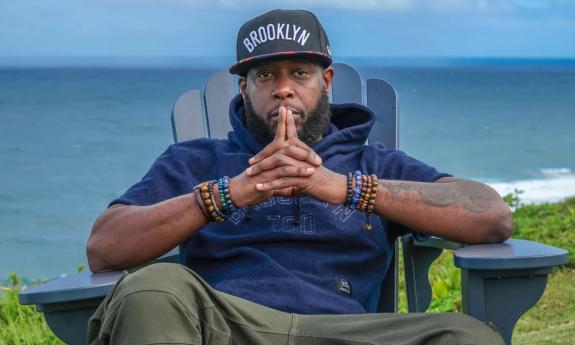 Talib Kweli sits back in a chair musing in front of an open ocean. 