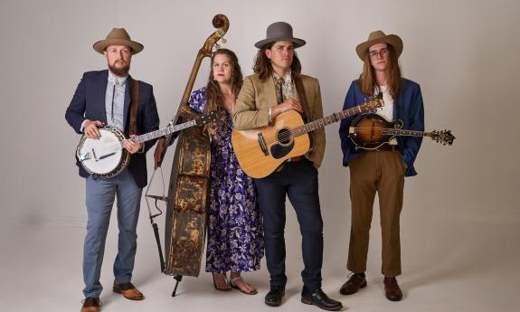 The four-person band, Remedy Tree, with their instruments, standing against a beige background