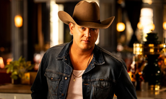 Jon Pardi poses with a brown cowboy hat and dark blue denim jacket in front of a bar with brown and amber hues. 