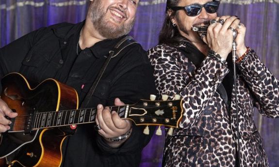 Nick Moss smiles while strumming his guitar on a stage with a purple backdrop.