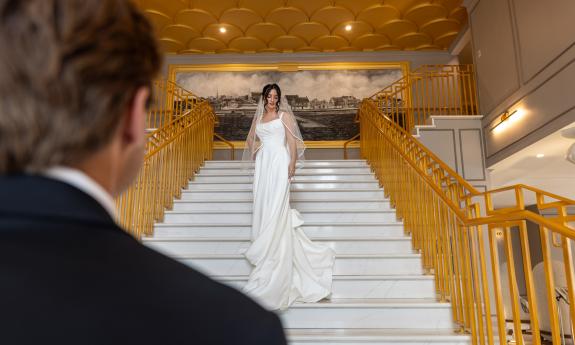 A bride on a white and gold stairway, with her groom looking up at her