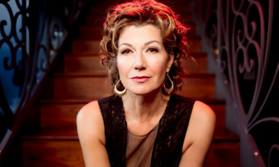 Amy Grant wears a black dress and poses in front of a brown staircase.