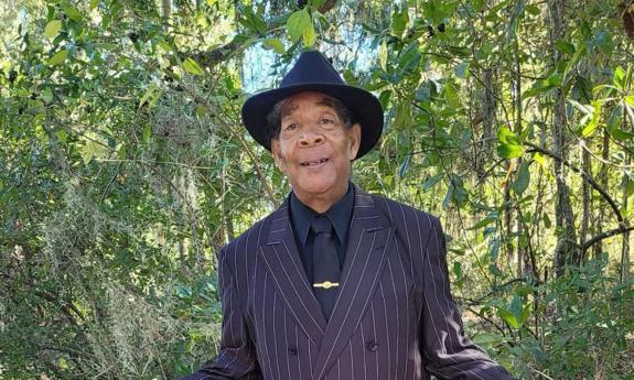 Bluesma Gitlo Lee in black hat, standing with woods behind him