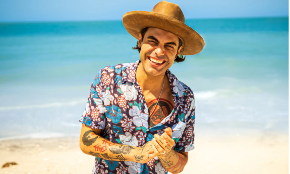 Niko Moon clasps his hands together and stands in front of a sandy beach.