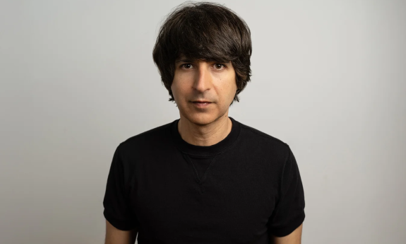 Demetri Martin dons a black shirt and poses in front of a gray backdrop.