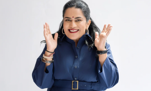 Zarna Garg stands before a white backdrop, beaming in a blue dress.
