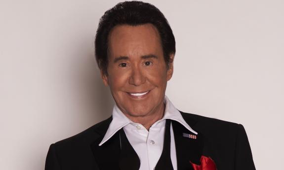 Wayne Newton wears a black suit while posing against a white backdrop.