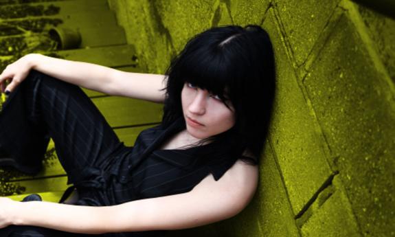 Patty PerShayla sitting in front of wall, looking up at camera. Image by Rae Jean Erickson