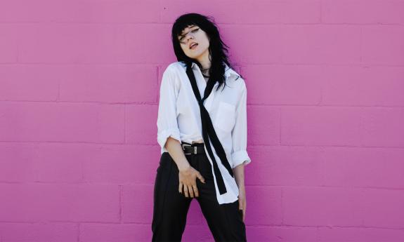 Patty PerShayla standing in front of pink wall in white blouse and black tie. Image by Rae Jean Erickson