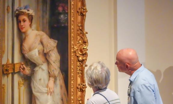 Two people look at a painting of a woman on display at the Lightner Museum