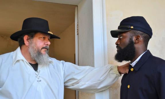 Two men, one older and white, and one younger and black dressed in clothing from the 1800s