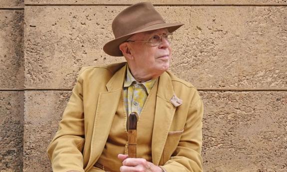 Bruce Cockburn poses while wearing a brown outfit and clutching a wooden cane.