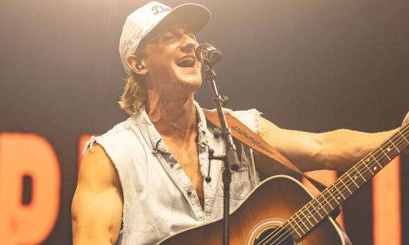 Cooper Alan wears a hat and strums his guitar on stage. 