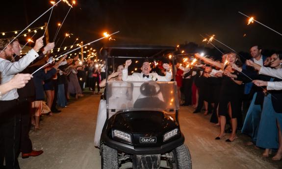 A bridal couple in a black golf cart, laugh as they pass guests with lighted wands