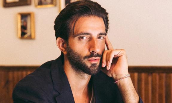 Davide De Pierro wears a black shirt and poses in front of a beige and brown wall. 
