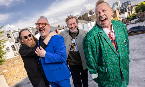 Members of Public Image Ltd. smile as they pose together against a wide, scenic neighborhood backdrop.