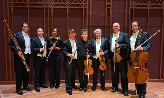 Eight musicians in a winds and strings ensemble, standing in a room with majestic wood paneling