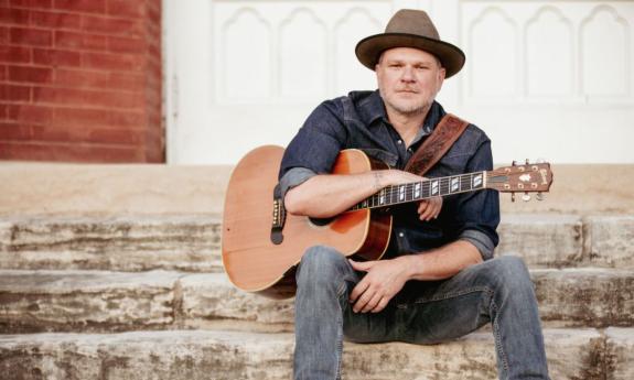 Jason Eady sits on a staircase and poses with his guitar.
